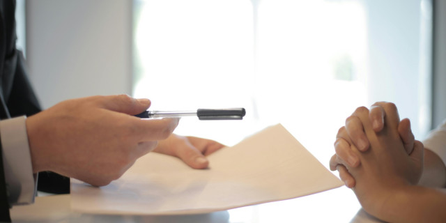 closeup of person in suit holding a paper and a pen opposite a person with folded hands.